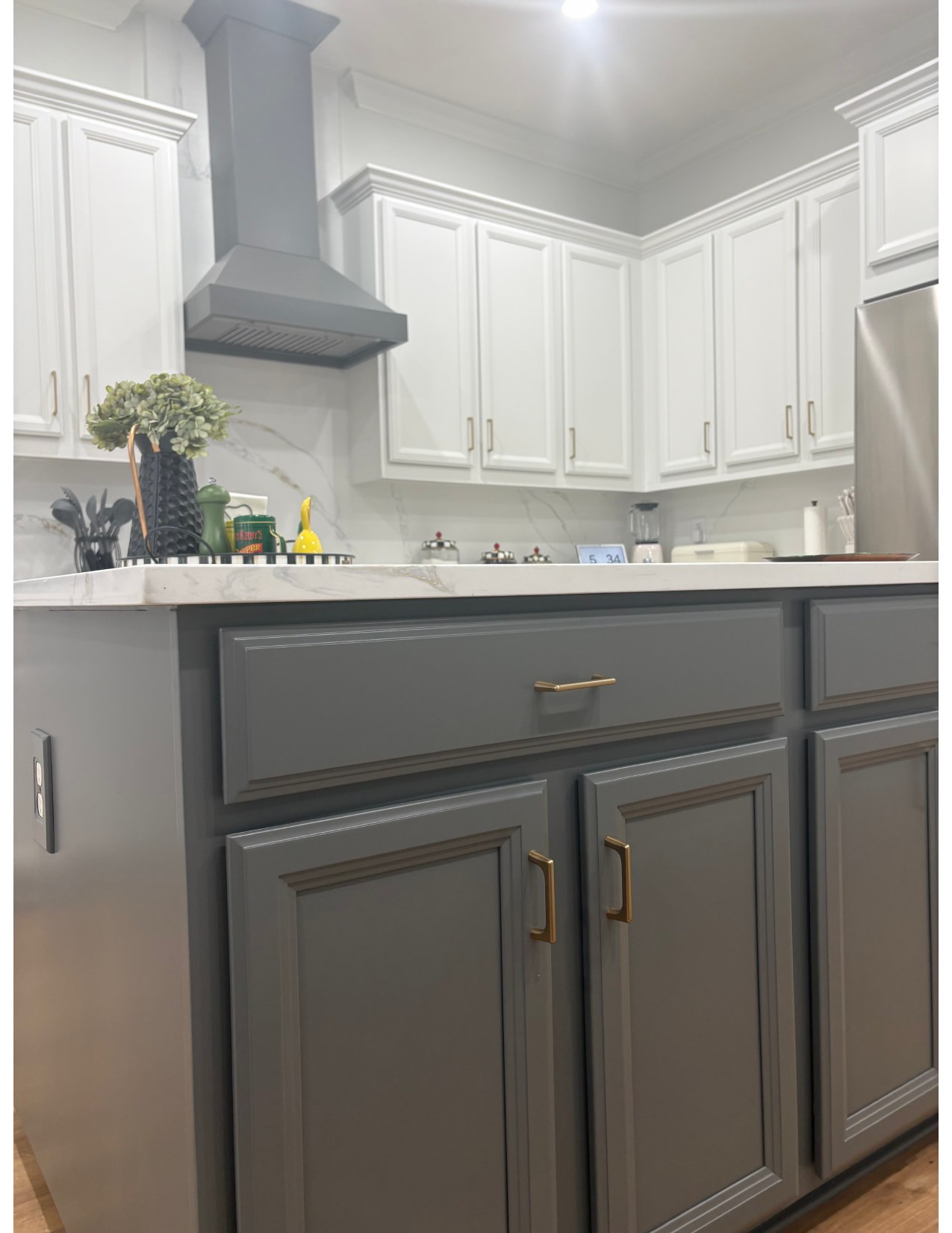 A two-tone refinished kitchen with white upper cabinets, charcoal-grey lower cabinets, brass hardware, and marble countertops