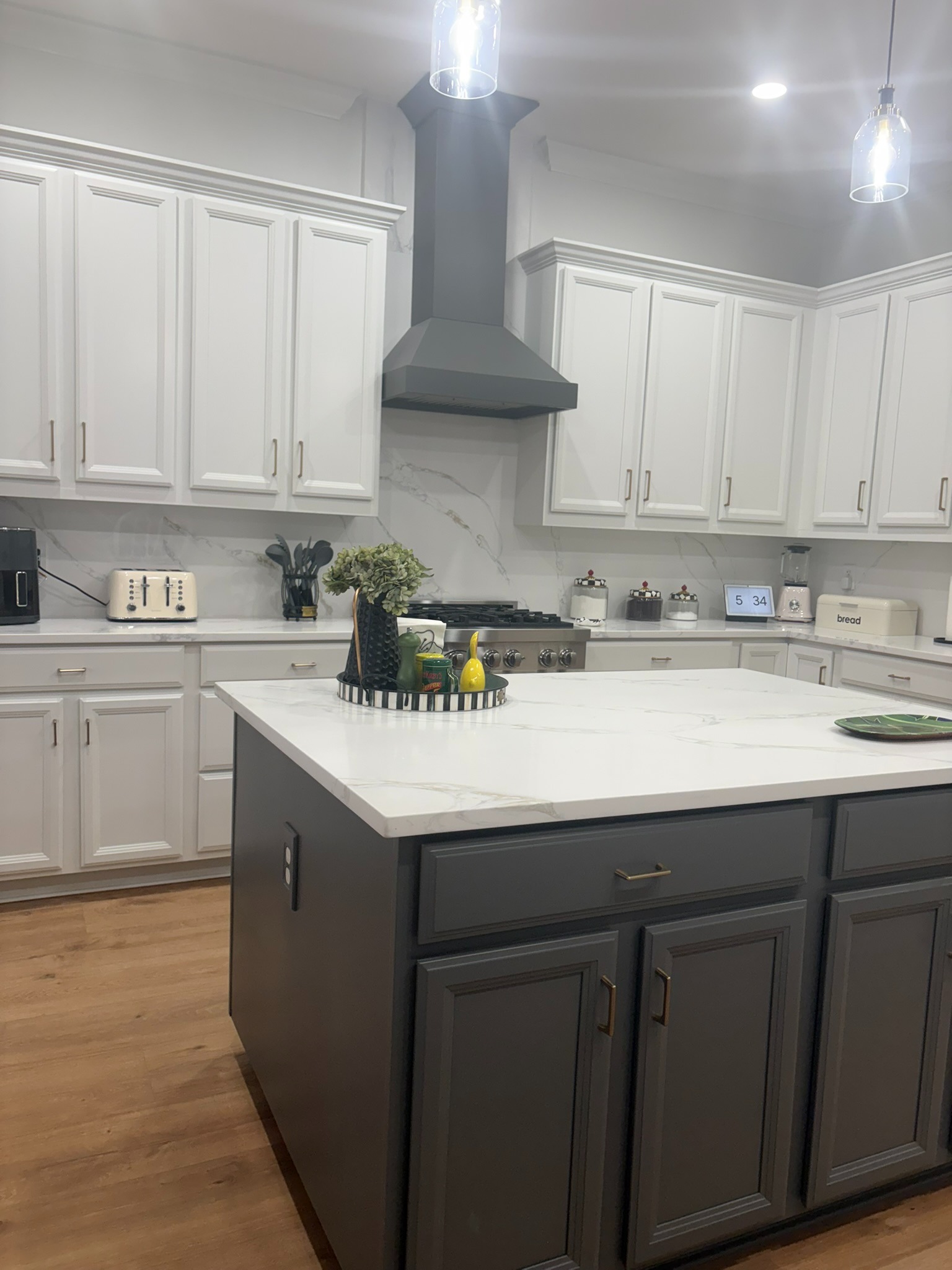 Wide view of two-tone refinished kitchen with white upper cabinets, charcoal-grey island and lower cabinets, brass hardware, and marble waterfall countertop